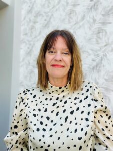 A picture of bid writer Karen Sherwood of George and Ivy Bid Writing - Karen has bob length brunette hair, is wearing a dalmation spotted dress, and stands in front of a white textured, outdoor wall.